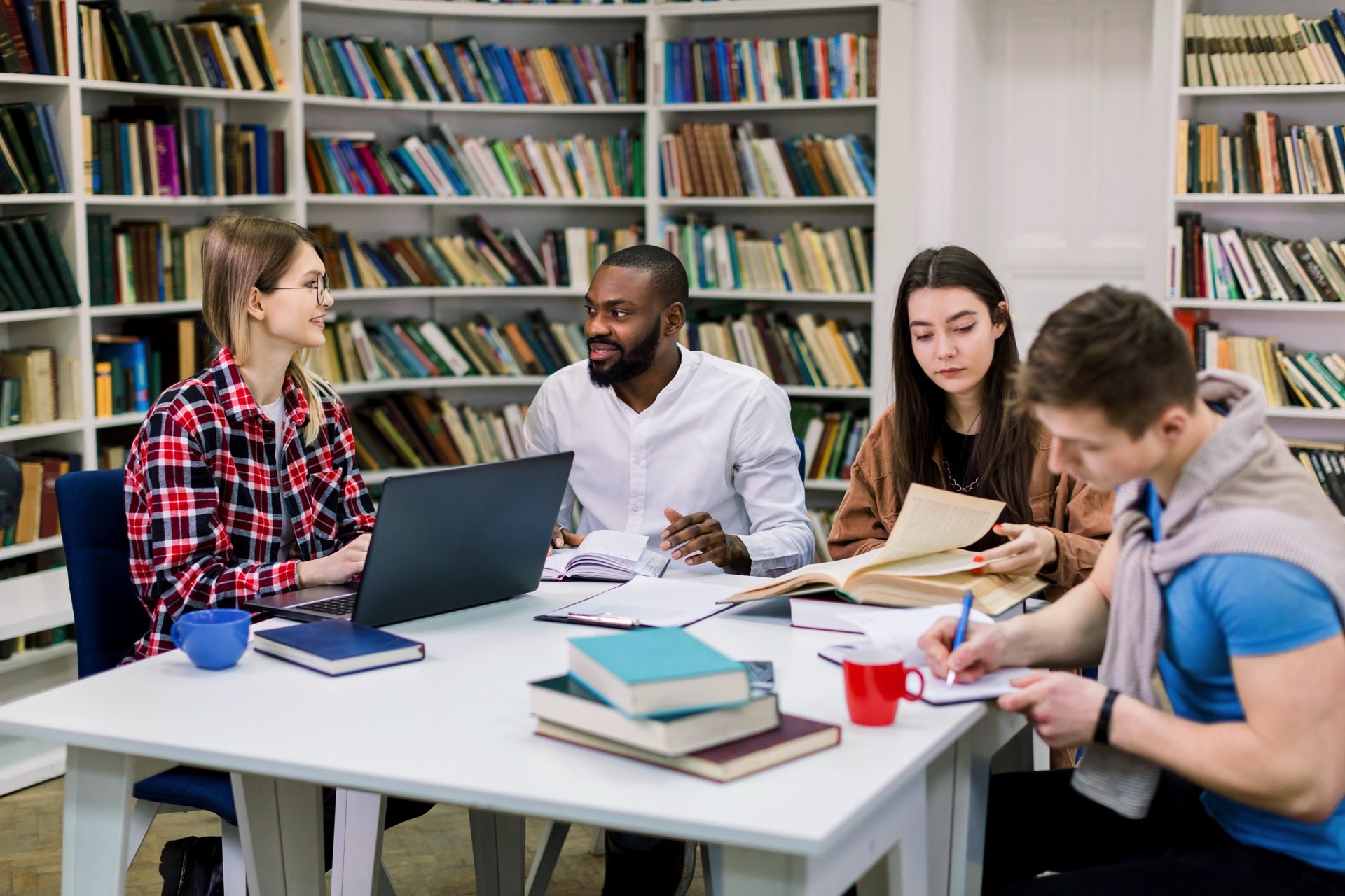 Four multiracial happy students sitting at table in library while learning and working on a laptop. Smart students which working together in the reading room using books and laptop Scots English Premises